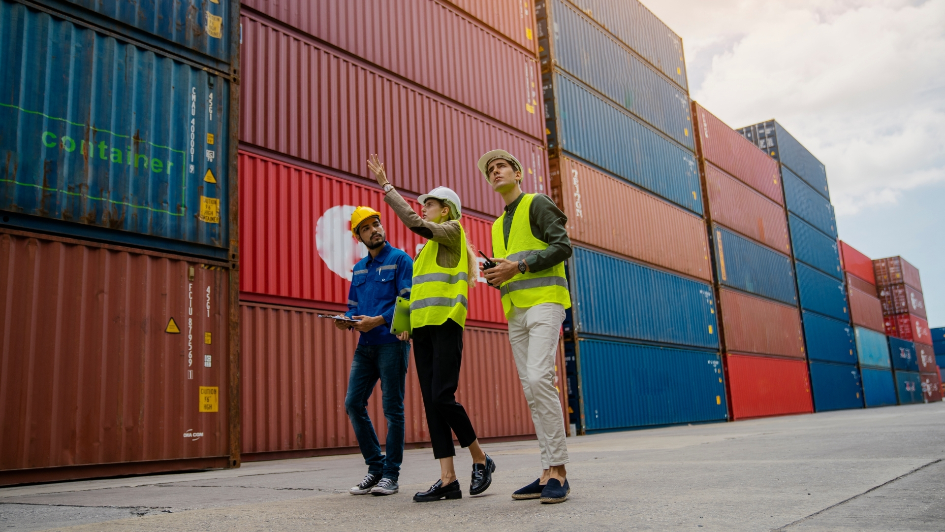 Three professionals in safety vests and helmets discussing logistics at a shipping container yard, with colorful containers stacked in the background.